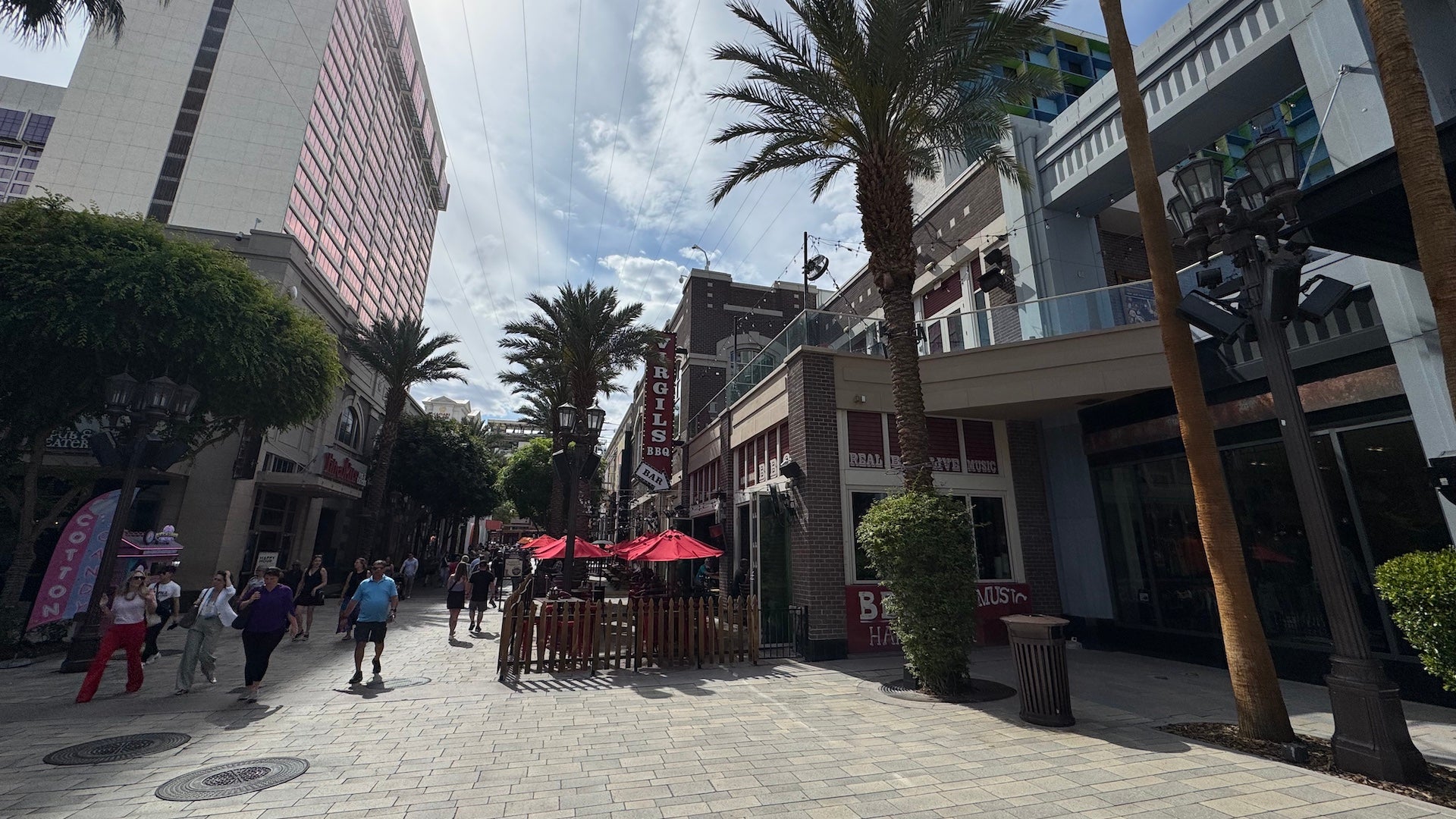 Pedestrian street scene at The LINQ Promenade in Las Vegas, featuring palm trees, outdoor diners under red umbrellas at Virgil’s Real BBQ, and surrounding shops and high-rise buildings under a partly cloudy sky.