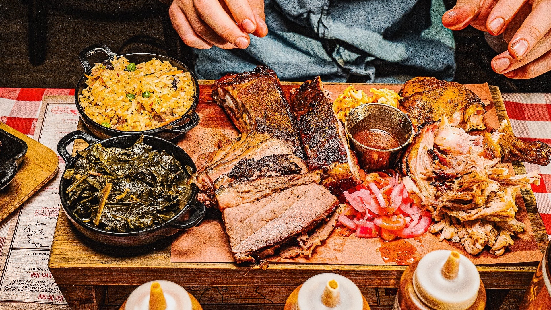 Close-up view of a hearty barbecue platter at Virgil’s BBQ, showcasing smoked brisket, ribs, pulled pork, barbecue chicken, collard greens, dirty rice, and pickled vegetables, with a small cup of barbecue sauce and squeeze bottles in the foreground.