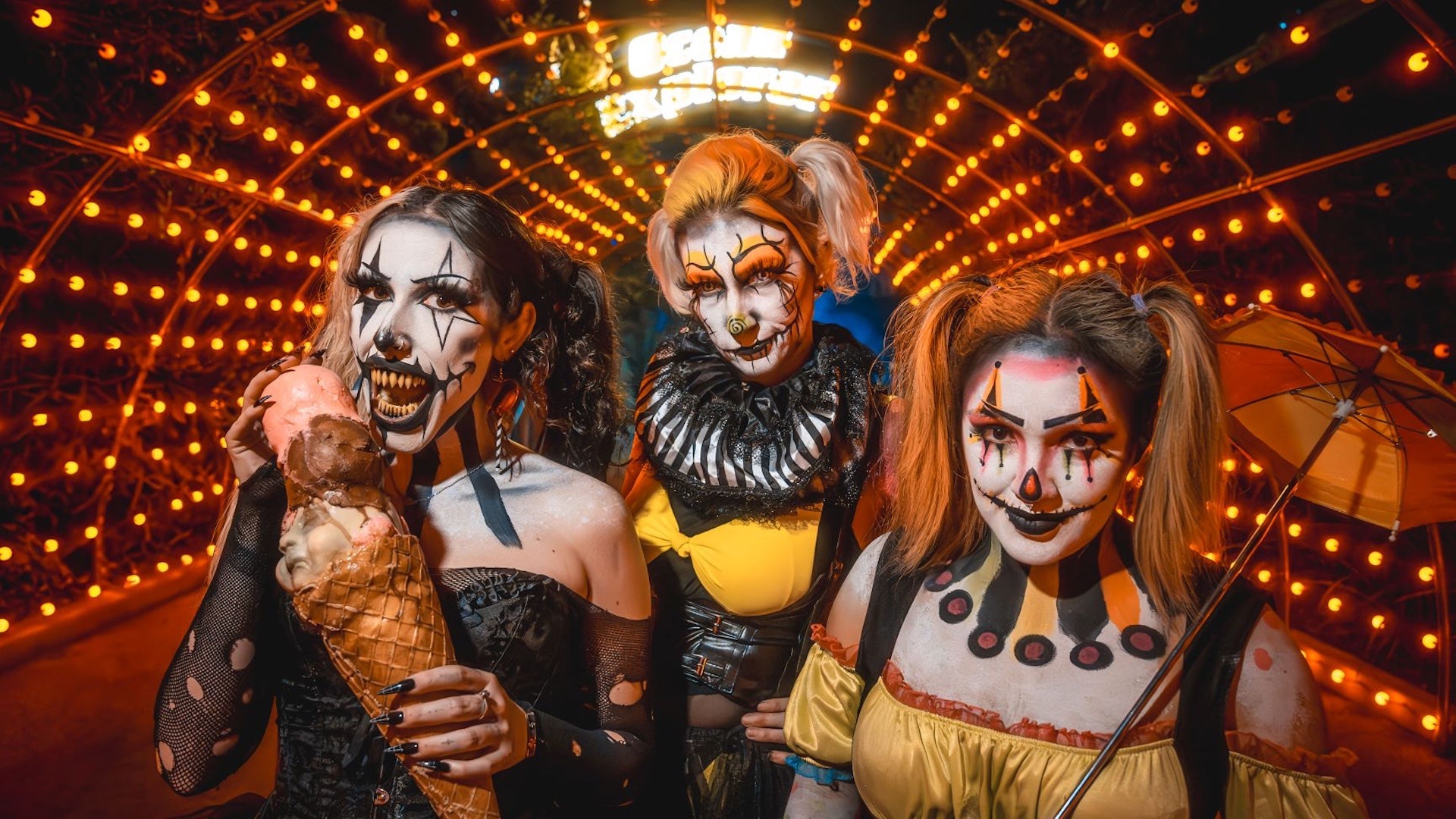 Three sinister clowns pose under an orange-lit tunnel. One bites into a grotesque ice cream cone with baby doll heads, another wears yellow with a ruffled collar, and the third carries a yellow parasol. All wear haunting makeup with exaggerated features, evoking a twisted circus vibe.