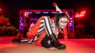 A creepy clown in black-and-white striped pants, a red ruffled costume, and a pointy hat crouches menacingly toward the camera under red stage lights at night. Her eerie makeup features blood-red eyes, sharp black-and-white contouring, and a sinister grin.