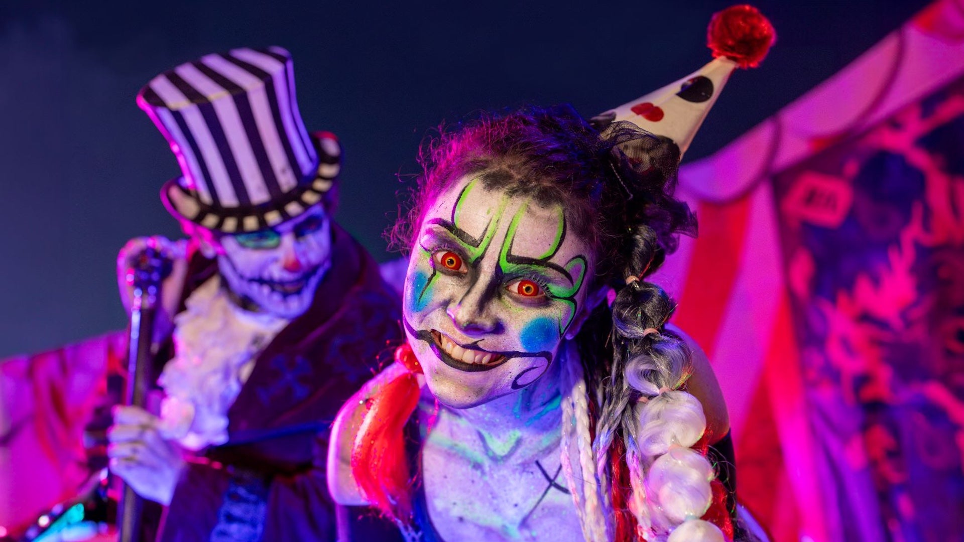 A glowing neon-painted clown with red eyes and green, black, and blue face paint leans toward the camera, grinning with intensity. She wears a party hat with hearts and pom-poms, while a menacing clown with a striped top hat looms behind her in a carnival-themed setting.