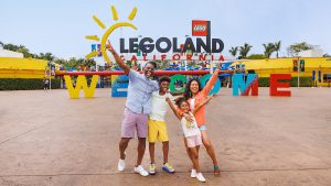 A smiling family of four poses with arms raised in front of the colorful “WELCOME” sign at the LEGOLAND California entrance on a sunny day.