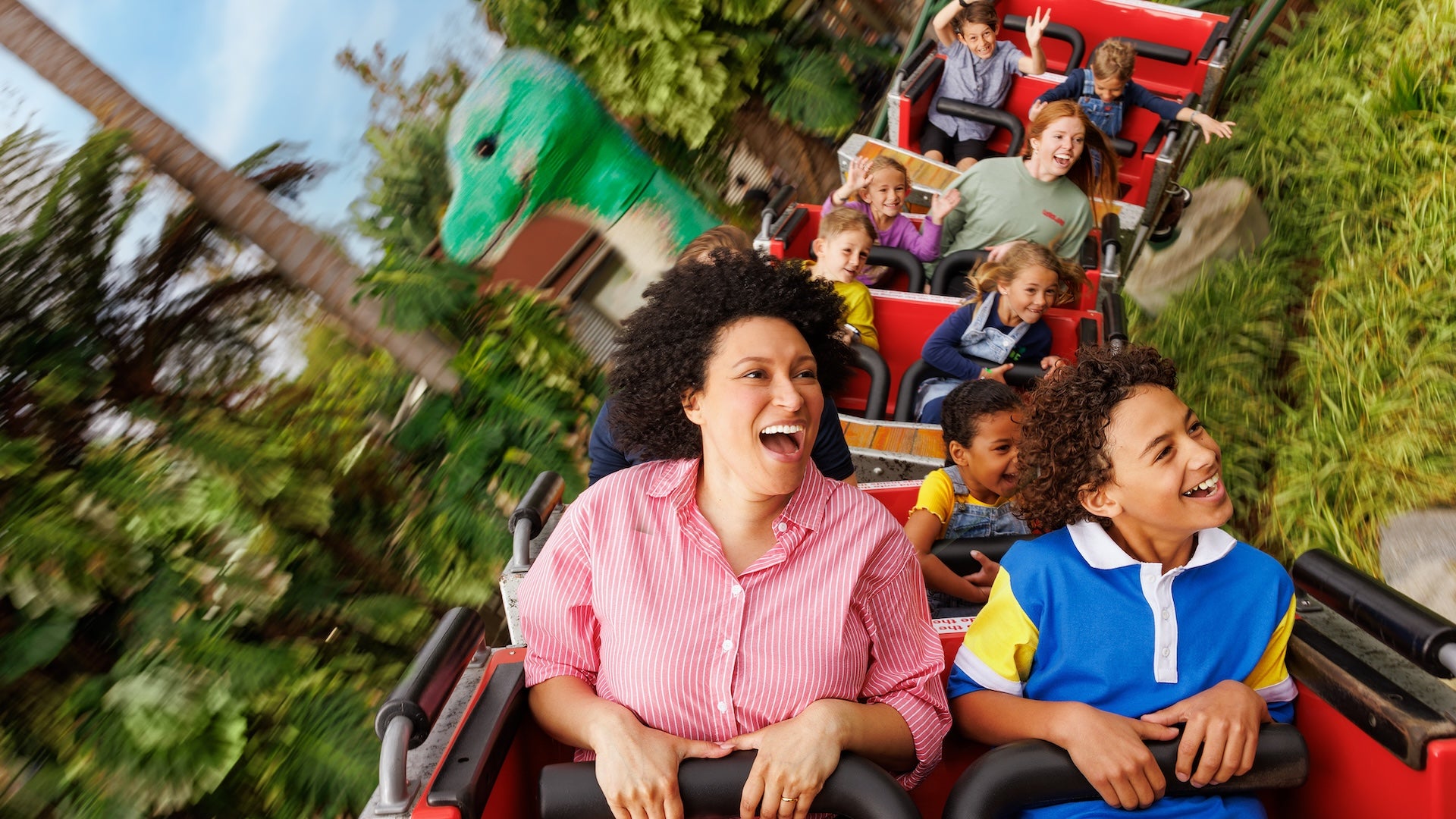 A joyful mother and son ride a red roller coaster at LEGOLAND California, with other excited passengers behind them and a large green LEGO dinosaur visible in the background.