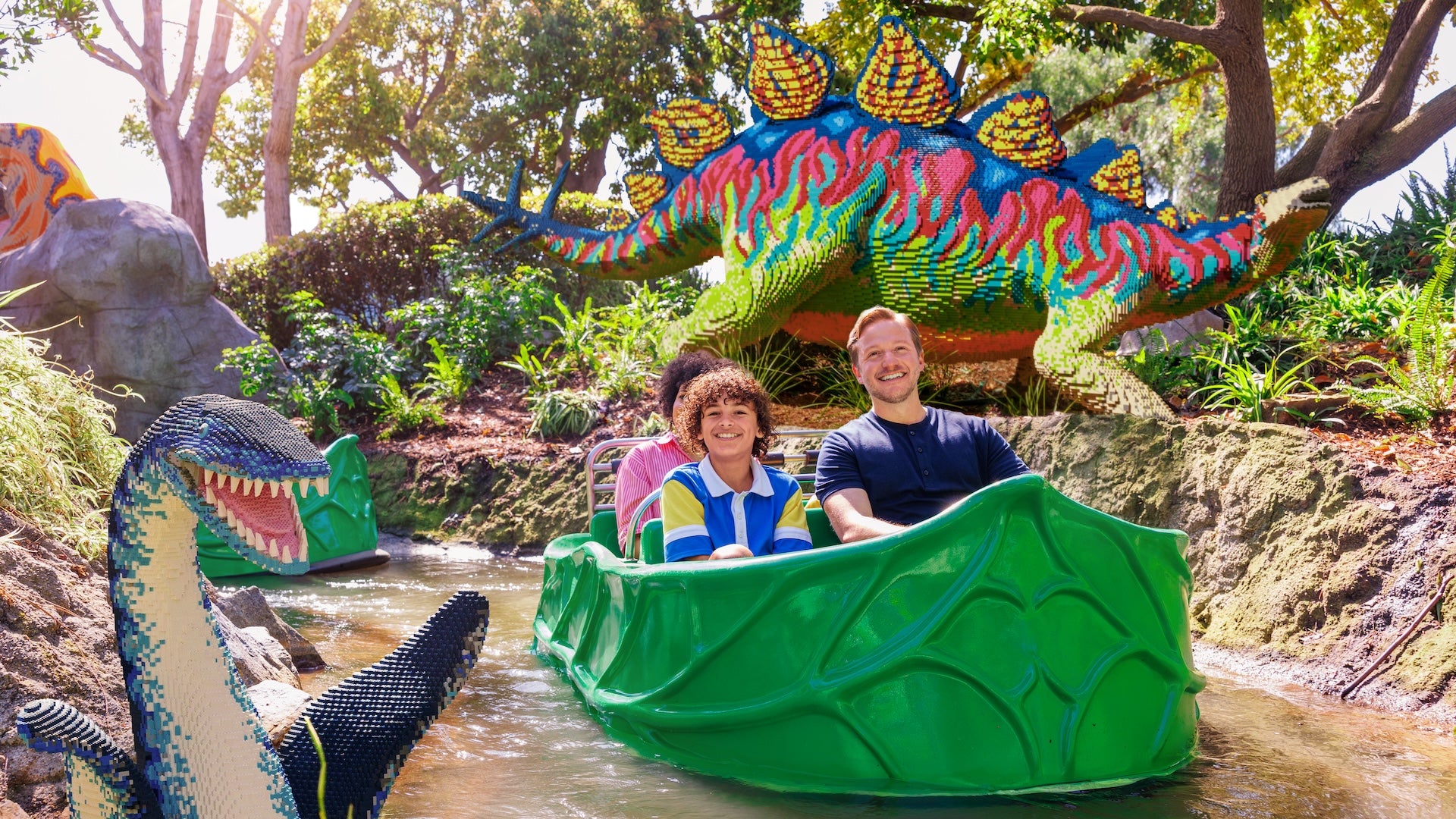 A father and son float in a green boat through Dino Valley at LEGOLAND California, passing by vibrant LEGO dinosaur sculptures surrounded by lush greenery.