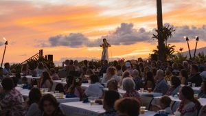 A woman dancing at a Luau with a sunset behind her and a crowd of people sitting at tables in front of her