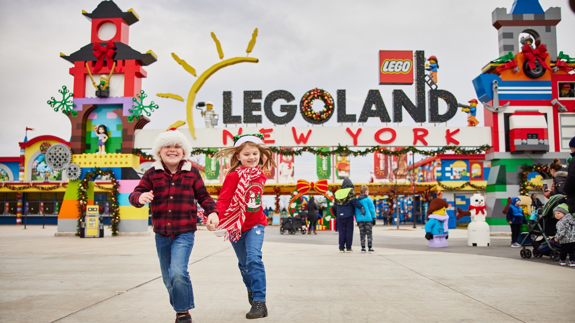 Two children holding hands running towards the camera with the entrance to LEGOLAND New York in the background