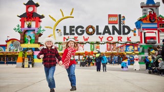 Two children holding hands running towards the camera with the entrance to LEGOLAND New York in the background