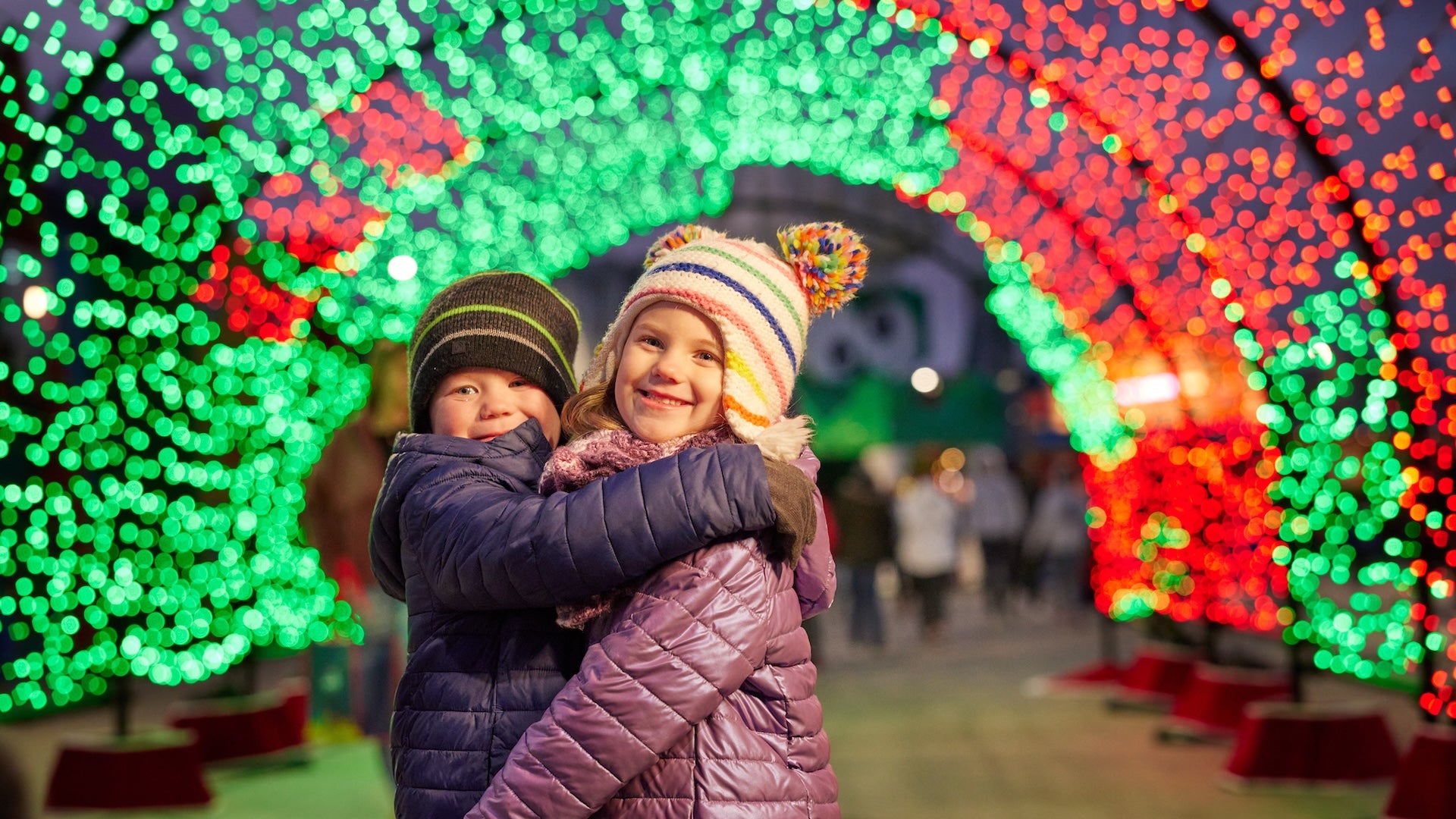 Two children in winter coats under an arch of Christmas lights at night