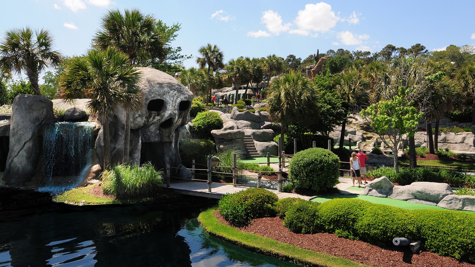 a couple on a mini golf course that has a skull cave and waterfalls, surrounded by trees