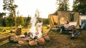 a girl camping with a fire pit in front of her and chairs beside her