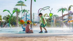 two kids playing on a water fountain in a water park with slides at the back and palm trees