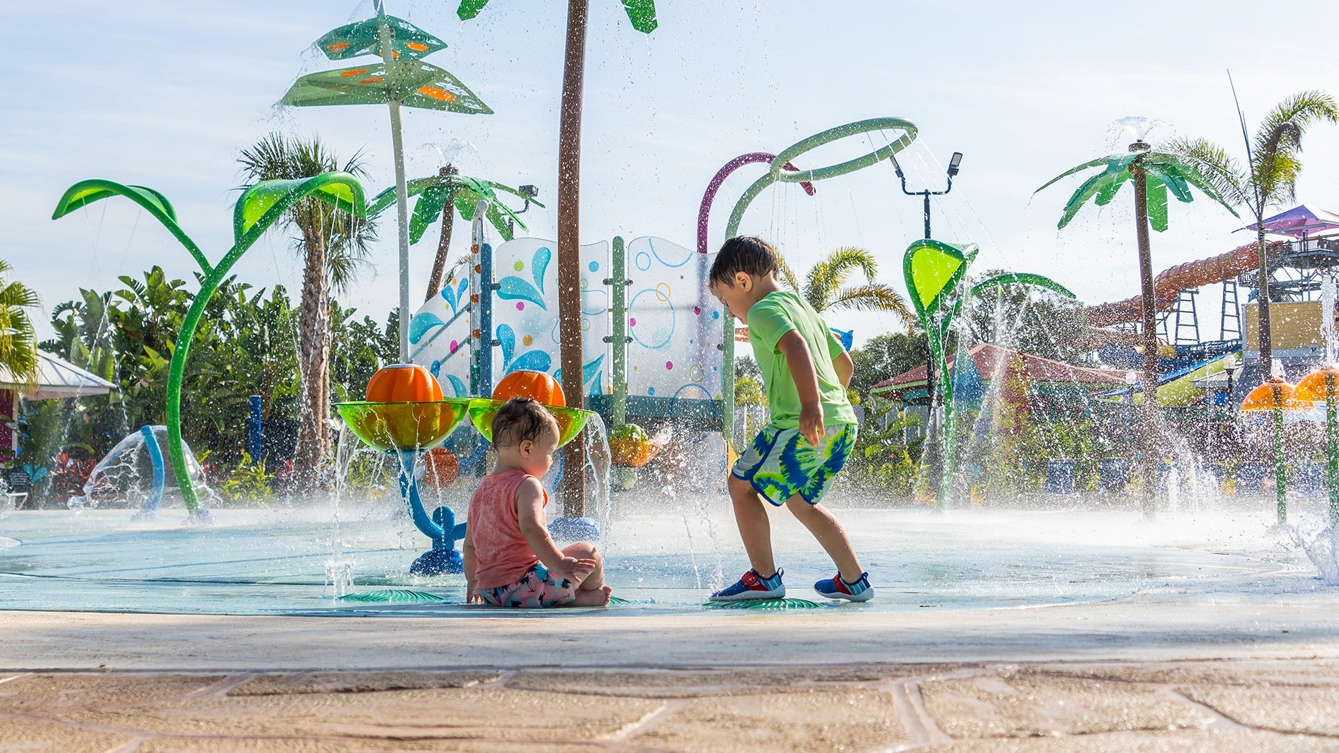 two kids playing on a water fountain in a water park with slides at the back and palm trees