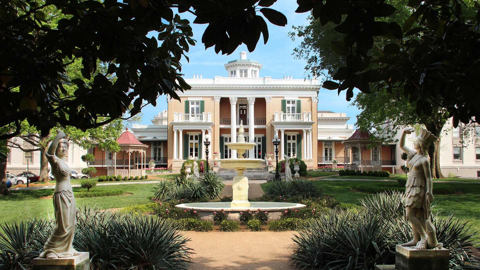 a mansion with a fountain in the middle of the garden, with two statues at the trail entrance