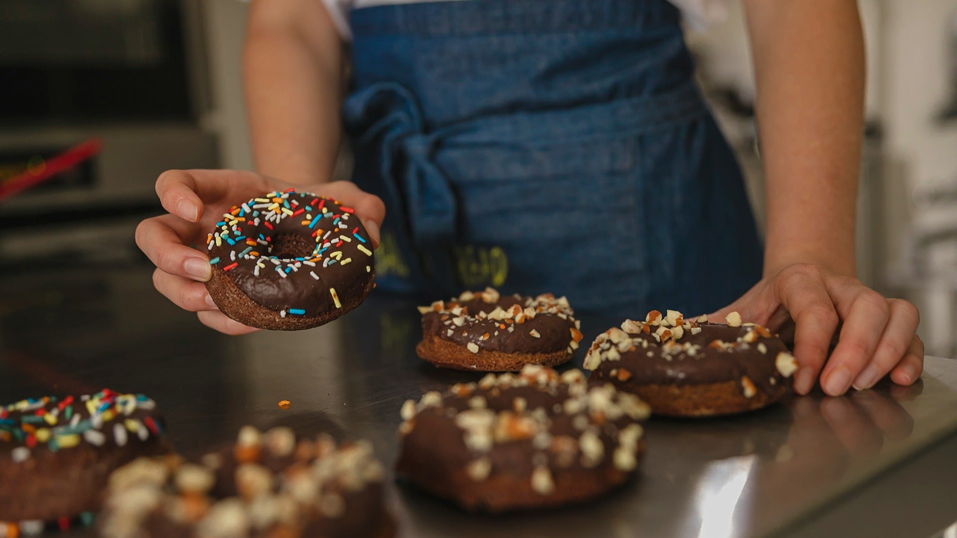 a woman holding a chocolate donut with sprinkles, more donuts on the table