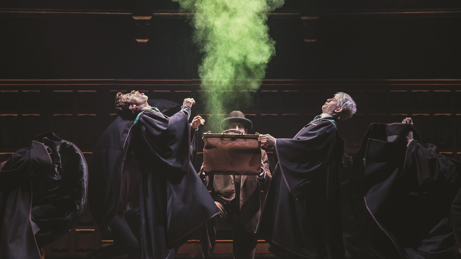 stage actors wearing cloaks and holding a bag with green smoke coming out of it