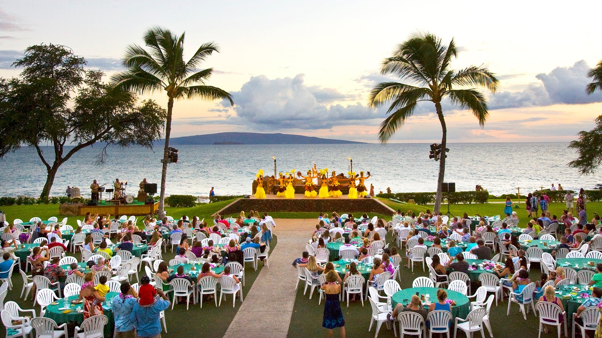 a luau with performers and a big audience and a view of the ocean behind
