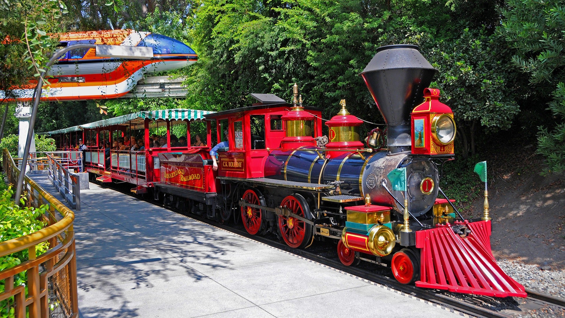 red steam train with passengers passing by trees