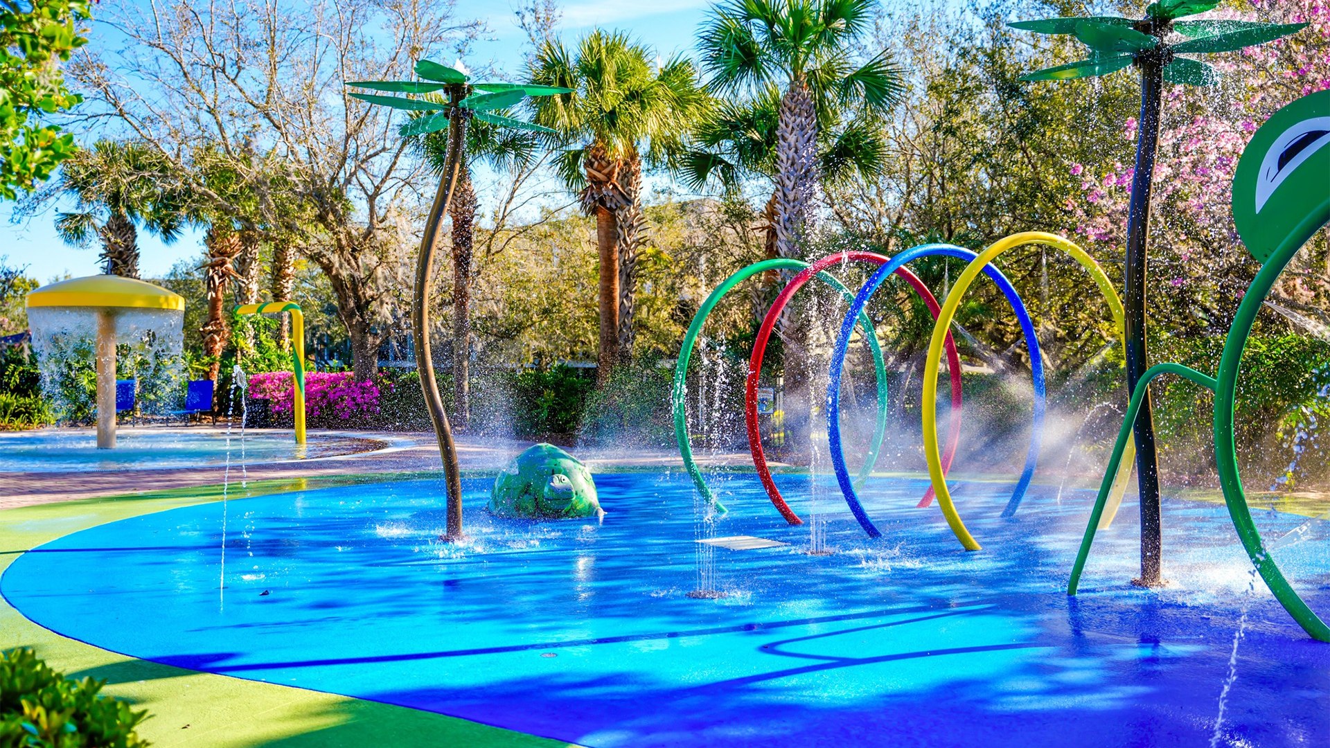 a brightly colored splash pad with rings and fountains and trees at the back