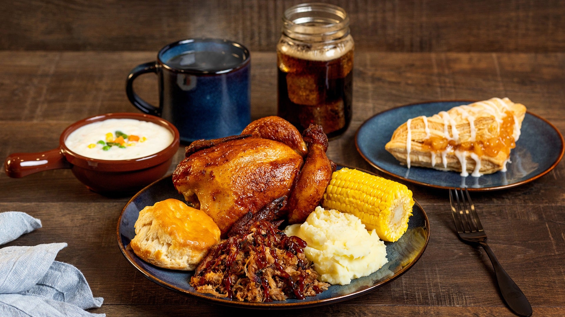 a food spread of roasted chicken, potatoes, corn, pies, soup, and drinks with a fork