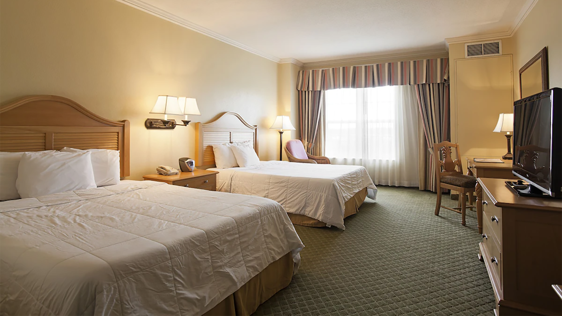 a hotel room with two beds, chair, lamps, tv, and big windows with curtains
