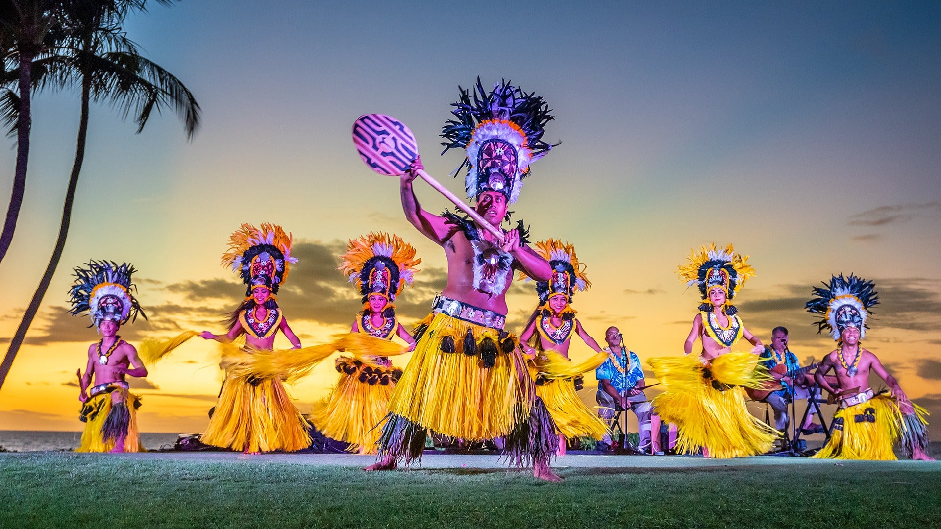 luau performers on stage, with dancers in costumes and singers at back while sunset