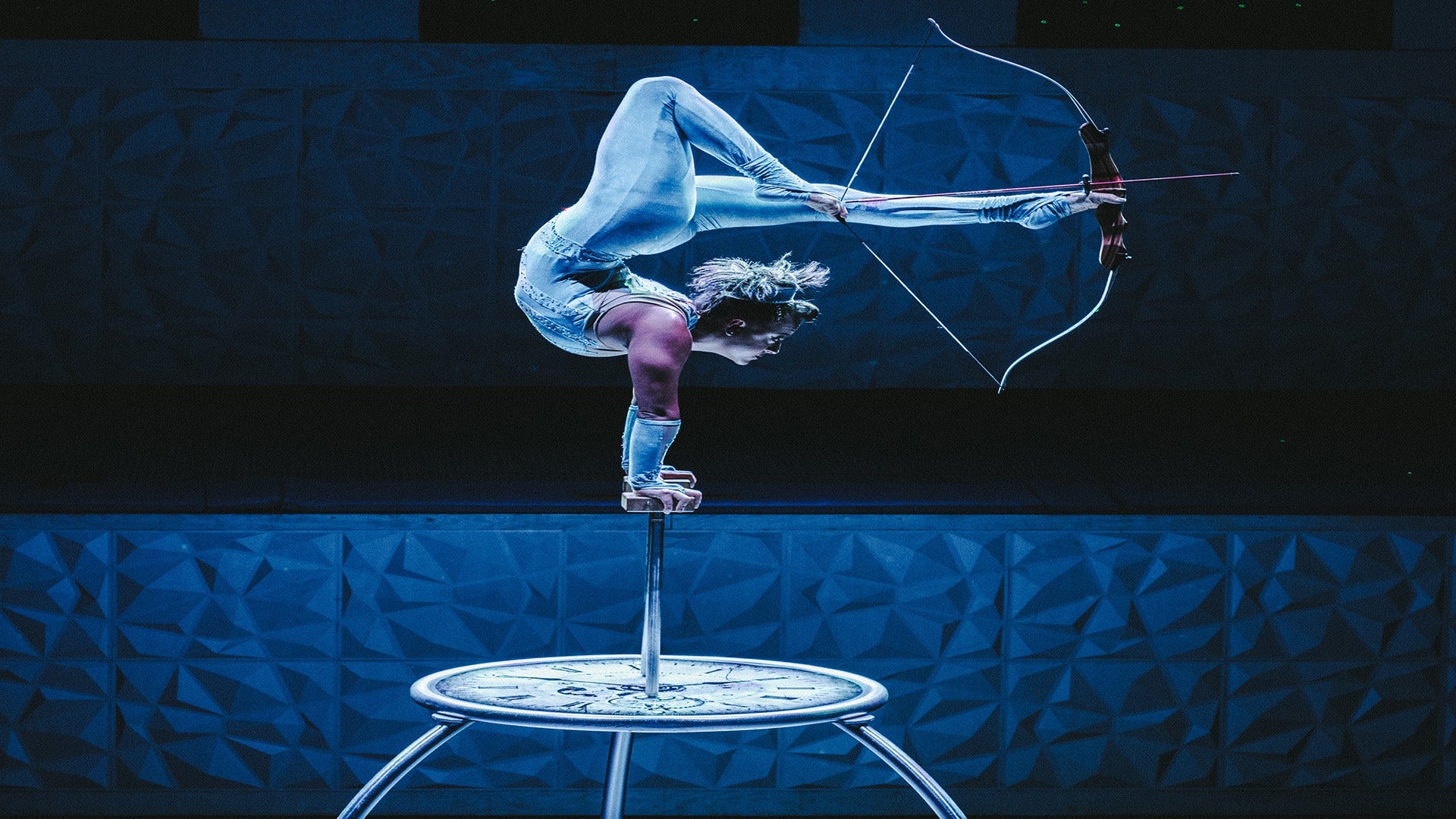 an acrobat shooting an arrow with his feet while balancing on a pedestal