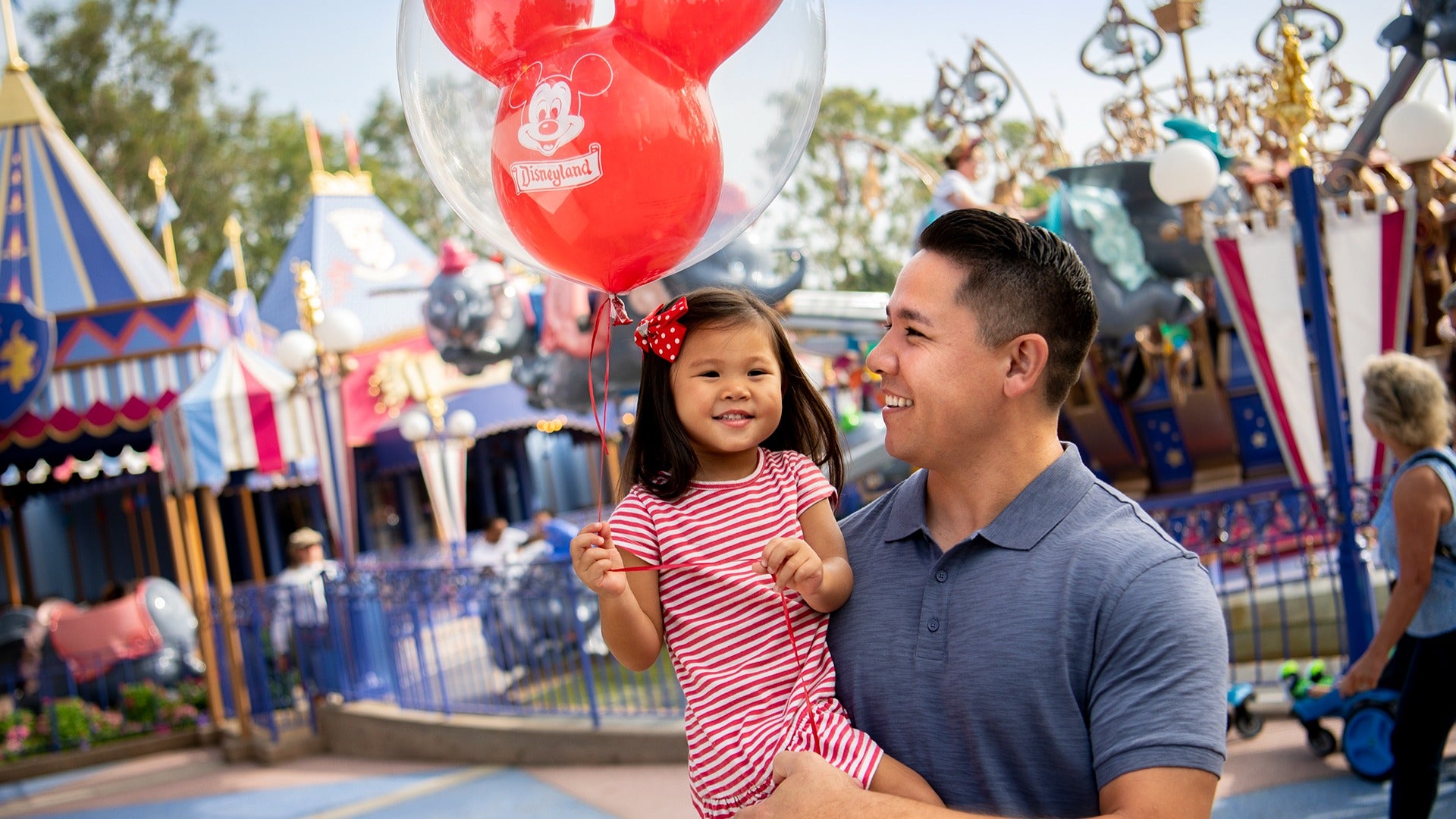 a father carrying his daughter holding a balloon at a theme park with rides behind them