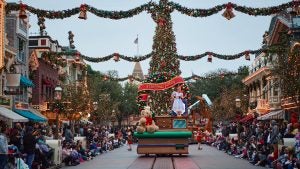 a holiday parade with a princess and a giant bear and a tree, with people on the streets watching it