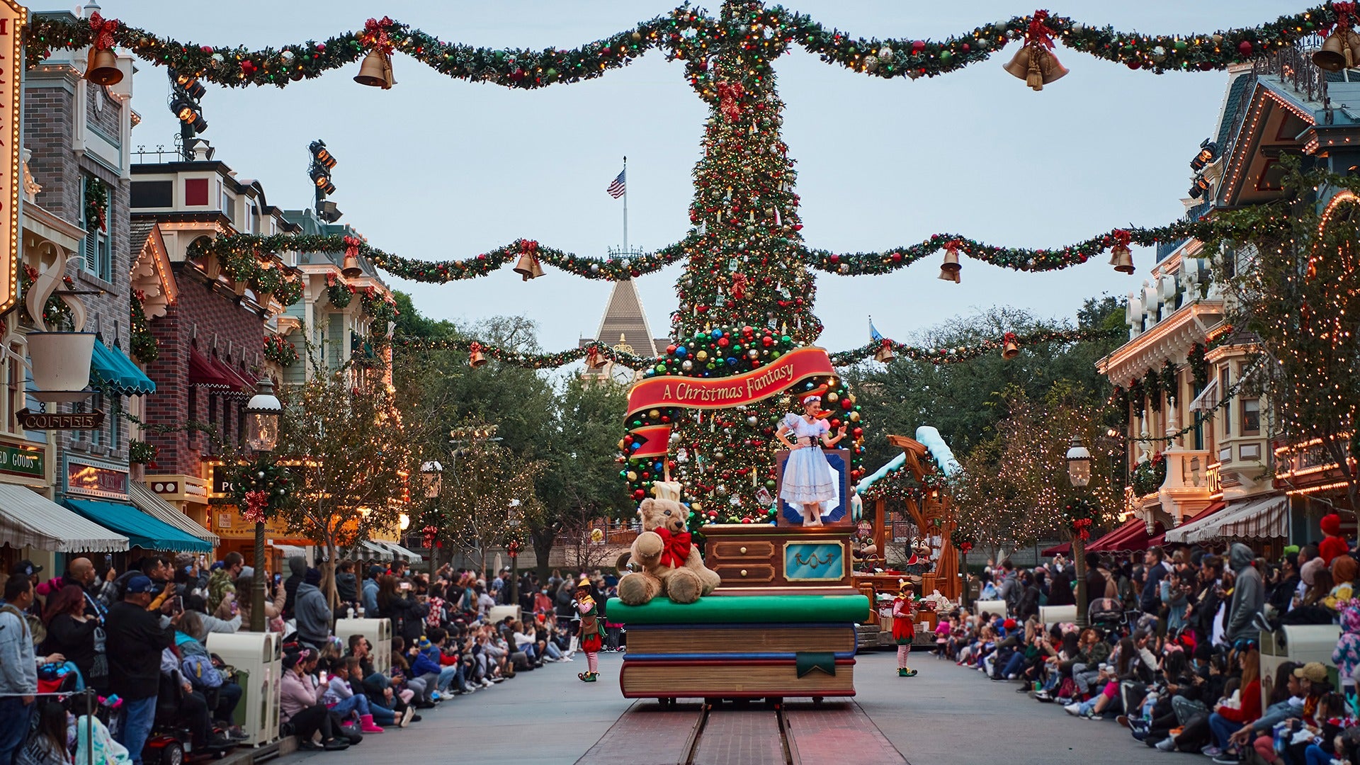 a holiday parade with a princess and a giant bear and a tree, with people on the streets watching it