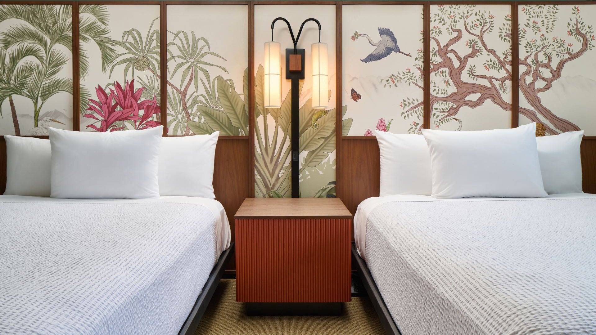 two hotel beds with a nightstand and lamps, wallpaper at the back