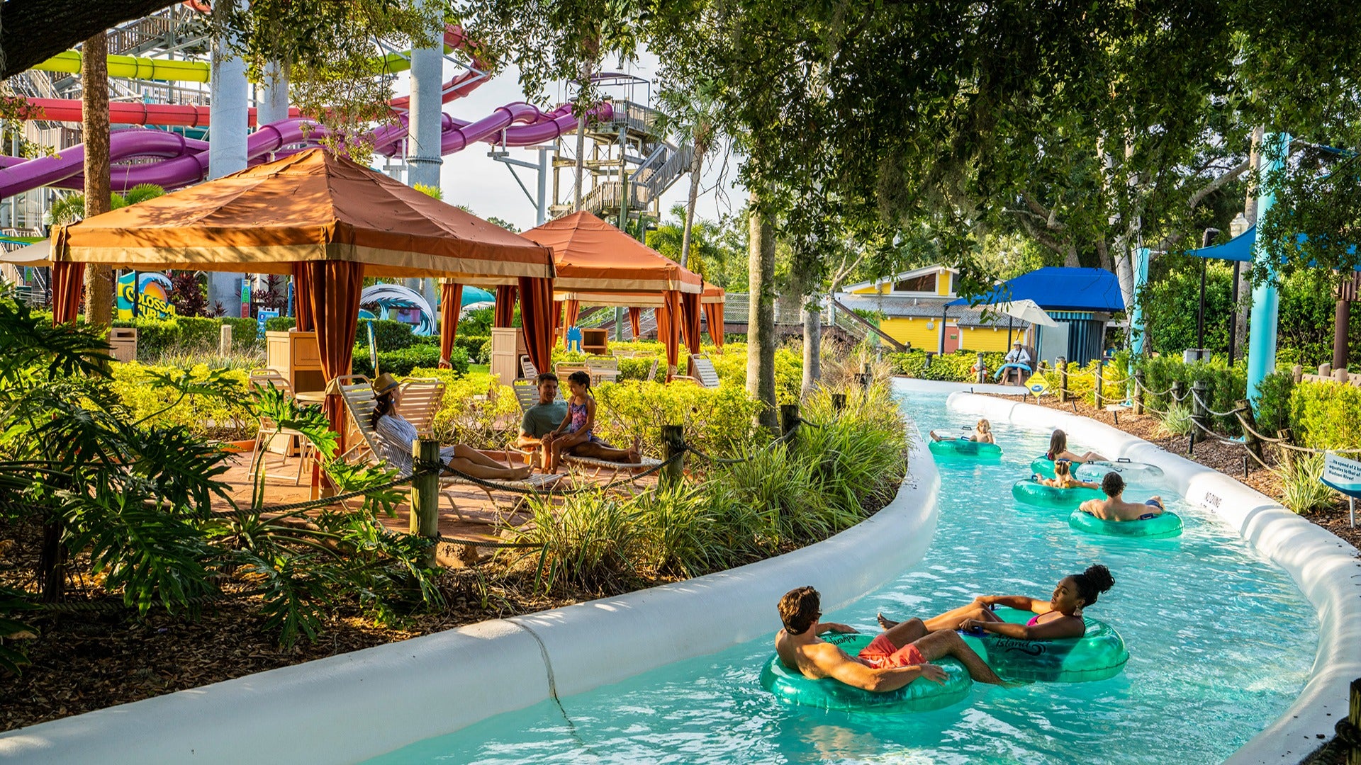 people floating down a lazy river with pool cabanas and chairs on the side, a water slide at the back