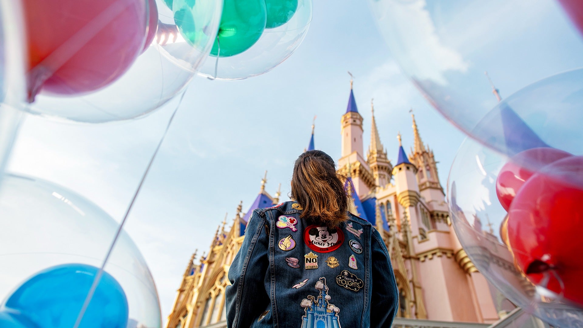 a girl wearing a disney patched jacket staring at cinderella's castle with balloons around her
