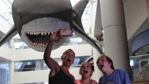 Family taking a selfie beneath a massive shark exhibit at the San Diego Natural History Museum, reacting with excitement and surprise