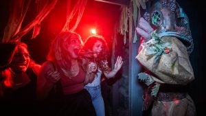 Three guests react with screams and laughter inside a dimly lit haunted house at Halloween Horror Nights Hollywood, coming face to face with a scare actor in a weathered deep-sea diving suit, bathed in dramatic red light