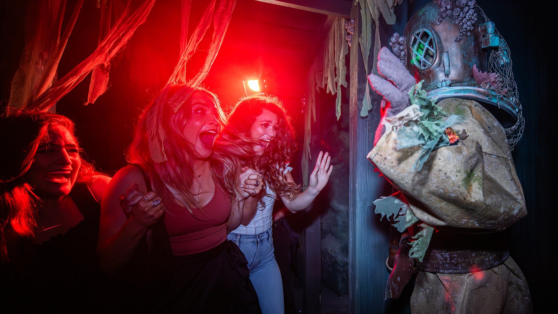 Three guests react with screams and laughter inside a dimly lit haunted house at Halloween Horror Nights Hollywood, coming face to face with a scare actor in a weathered deep-sea diving suit, bathed in dramatic red light