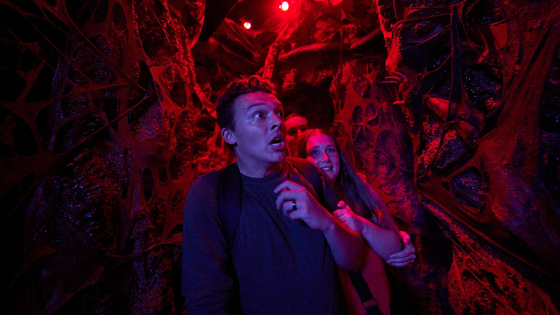 A man and woman navigate a narrow, red-lit corridor inside a haunted house at Halloween Horror Nights Hollywood, surrounded by grotesque organic walls as the man looks up wide-eyed with an expression of genuine unease