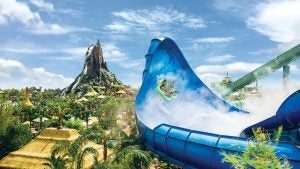 Guests ride a multi-person raft down a large blue water slide at Universal's Volcano Bay, with the park's iconic Krakatau volcano towering above a lush tropical landscape in the background