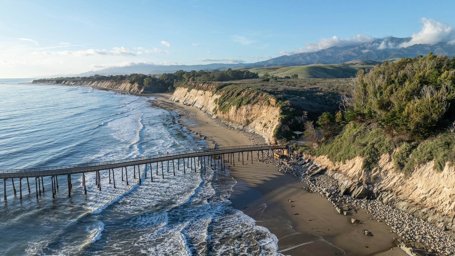 coastline with a pier going out into the ocean and mountains in the background under a blue sky