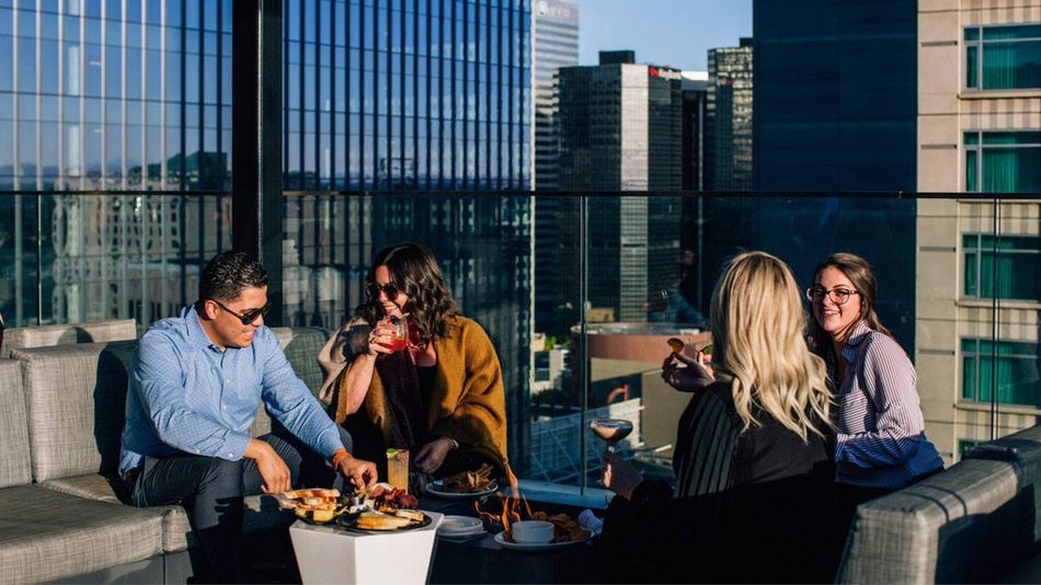 group of friends sitting on chairs with grey cushions having food and drinks on rooftop with view of nearby buildings during daytime at 54Thirty in Denver, Colorado, USA