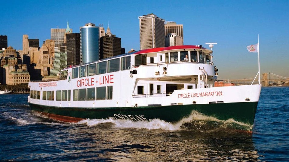 Circle Line Cruise on water with buildings and bridge in background during day with blue sky in New York City, New York, USA
