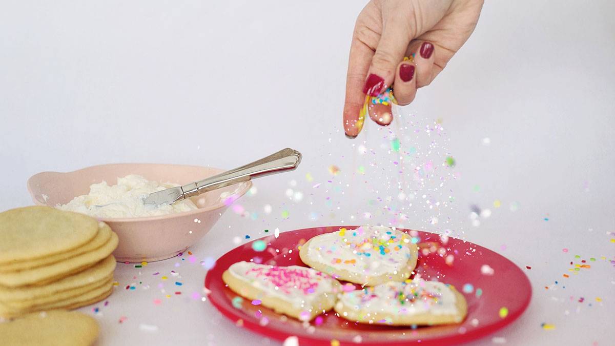 hand tossing colorful sprinkles and edible glitter on frosting covered cookies on a plate with a bowl of frosting and cookies on the side