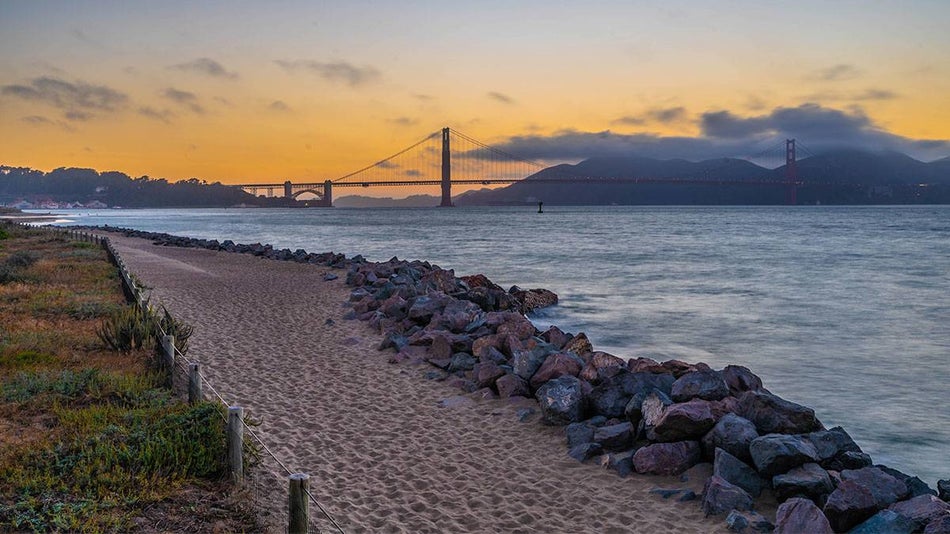 path with sand and rocks on the side with body of water and view of golden gate bridge during sunset at Crissy Field in San Francisco, California, USA
