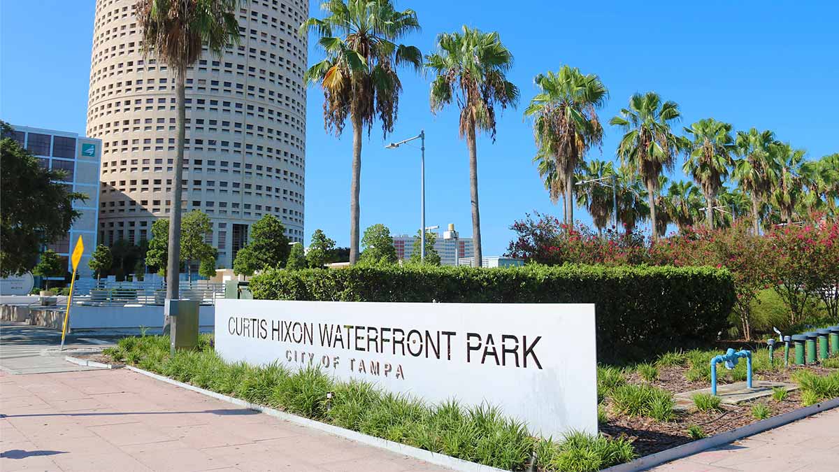 Curtis Hixon Waterfront Park sign with plants, row of palm trees and buildings in background during daytime in Tampa Bay, Florida, USA