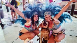 Two girls with blue feather crowns crouched down hugging a tan dog