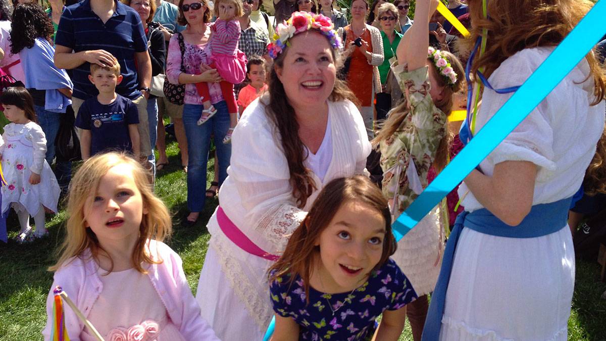 woman with flower crown wearing white holding blue ribbon with children dancing Maypole with crowd in background at Flower Mart in Washington National Cathedral, Washington DC, USA