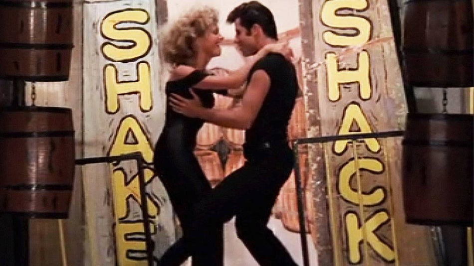 movie still from the movie Grease with two people dancing together with Shake Shack sign in background