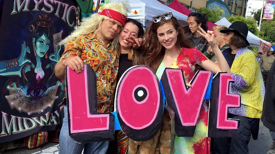 friends in tie-dye clothing posing for photo holding large LOVE sign with crowd in background at Haight Ashbury Street Fair in San Francisco, California, USA