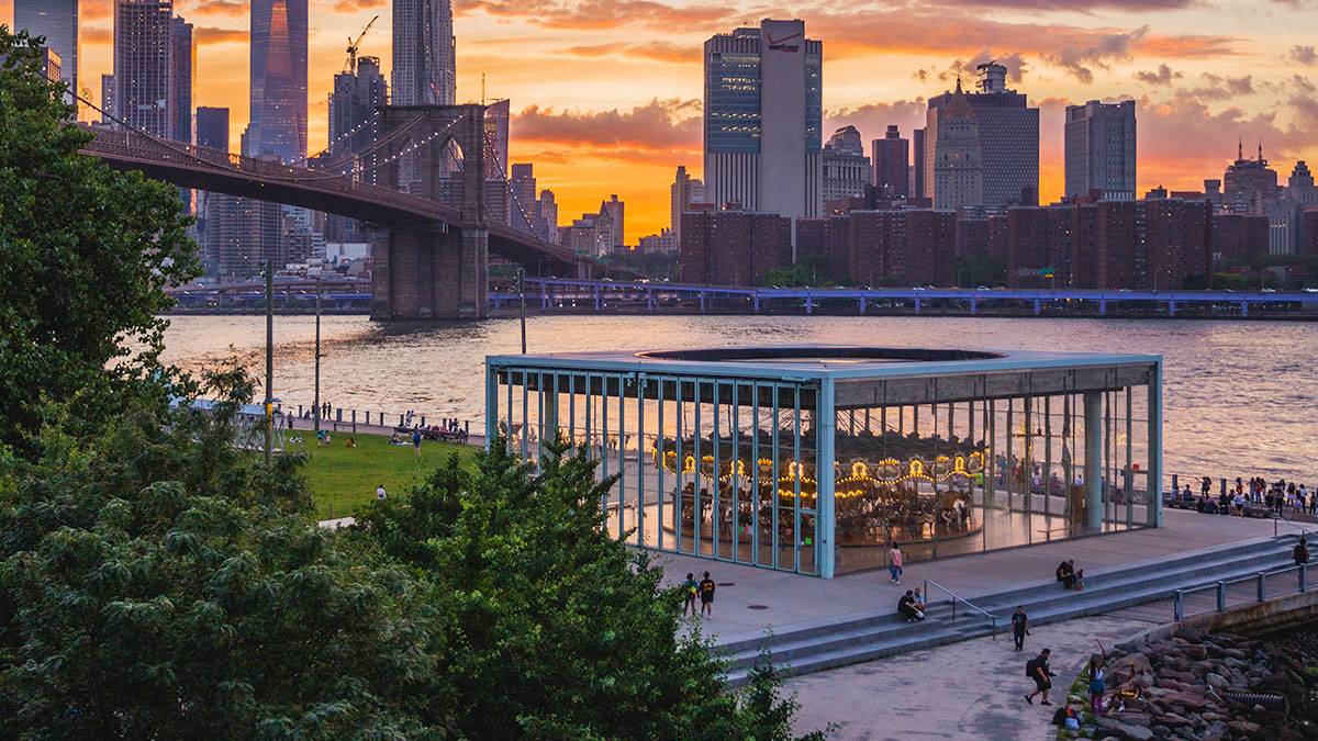 aerial view of Jane's Carousel with people and trees in the foreground and Brooklyn Bridge and buildings in background during sunset in New York City, New York, USA