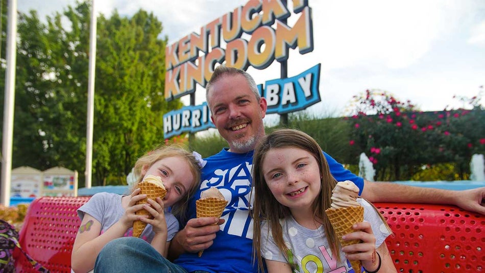 father with two daughters sitting on red bench eating icecream beside entrance of Kentucky Kingdom with large sign in Kentucky Kingdom, Louisville, Kentucky, USA