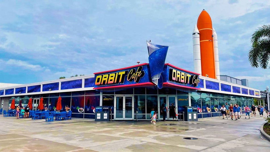 people walking beside exterior of Orbit Cafe with orange space shuttle attached to establishment at Kennedy Space Center in Orlando, Florida, USA
